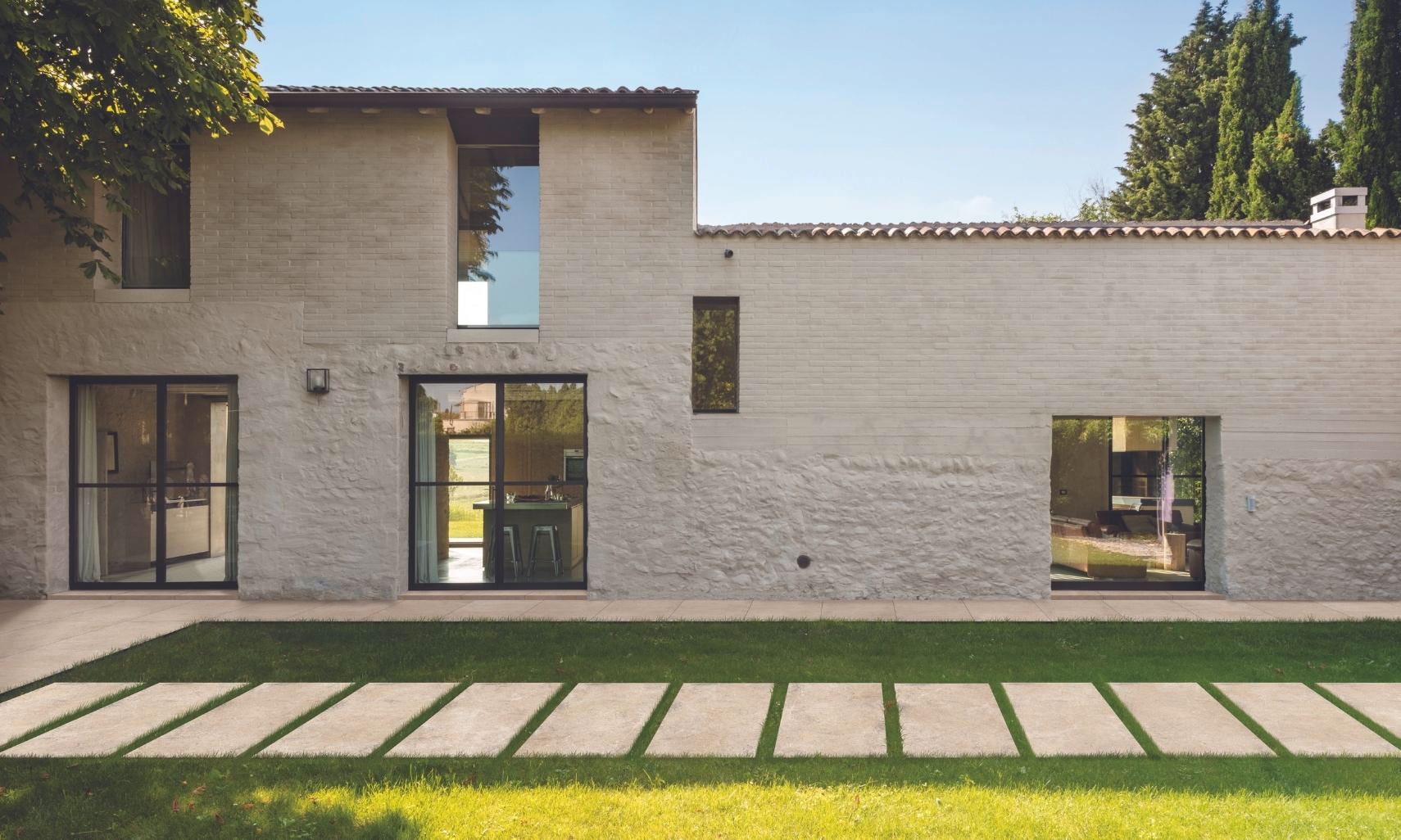 Large beige tile pavers that look like stone, set in grass in front of a beige Spanish style home with large picture windows and clay tile roof.
