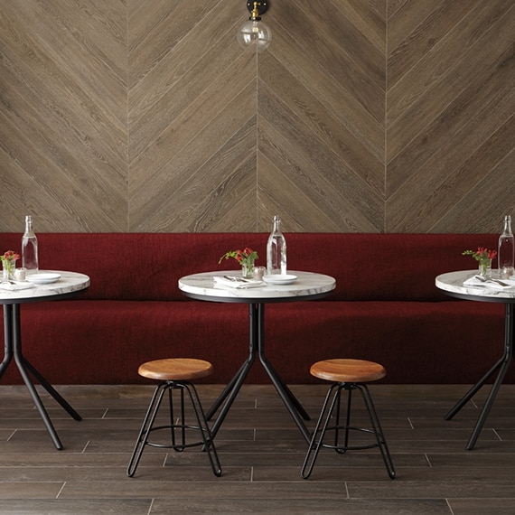 Restaurant dining room with wall-length booth covered in red velvet, herringbone wood look wall tile, marble top table, wood look tile flooring.