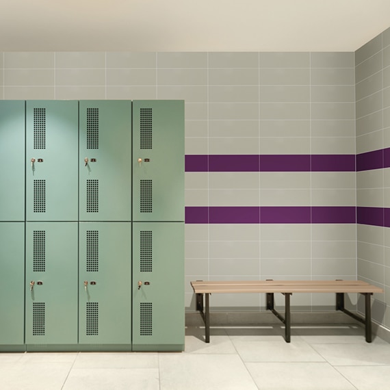 Locker room with 24x24 beige concrete look, beige wall tile with 2 purple horizontal stripes, mint green lockers and wood bench.