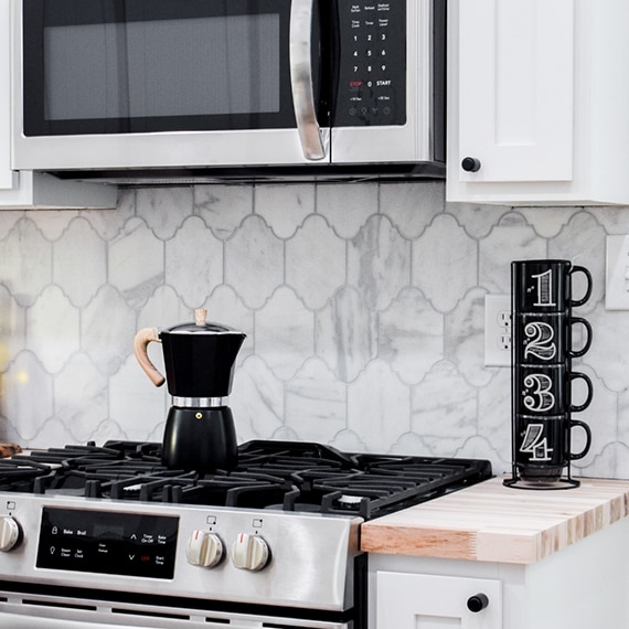 Kitchen backsplash of white & gray, Victorian mosaic marble tile, butcher block countertop, stainless steel gas stove and microwave.